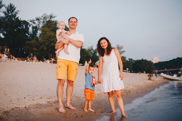 View Of Family With Two Toddler Children Outdoors By The River In Summer. Happy Young Family Have Fun On Beach At Sunset. Active parents and people outdoor activity on summer vacations with children