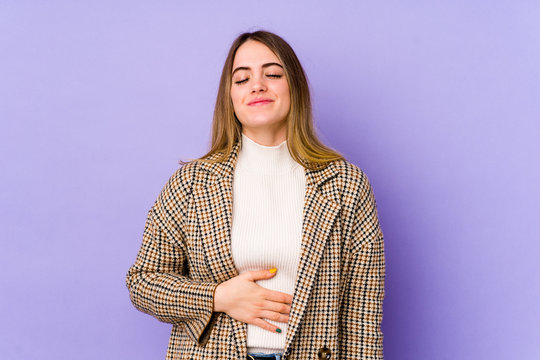 Young caucasian woman isolated on purple background touches tummy, smiles gently, eating and satisfaction concept.