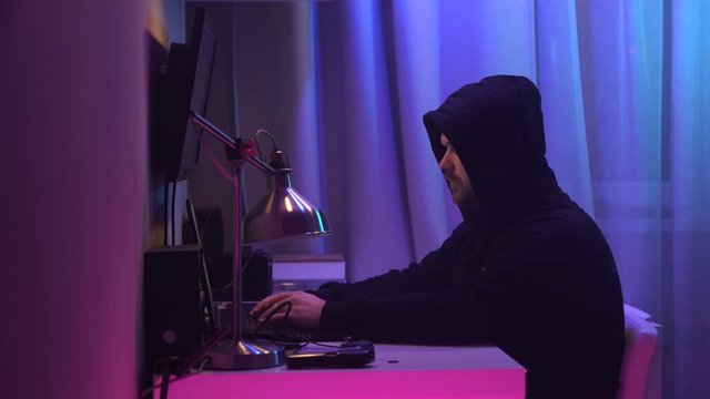 Male hacker sits at his desk and breaks into the system. Man in black hoodie hides his face and works at laptop at neon lighting.