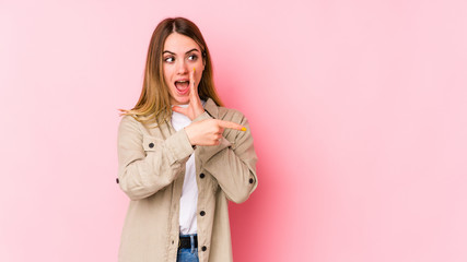 Young caucasian woman isolated on pink background saying a gossip, pointing to side reporting something.