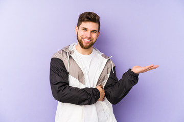 Young man isolated on purple background showing a copy space on a palm and holding another hand on waist.