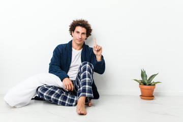 Mature man wearing pajama sitting on house floor showing number one with finger.