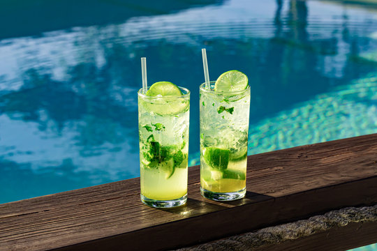 Two fresh and cool mojitos with ice cubes, lime slices and mint leaves on a turquoise pool background