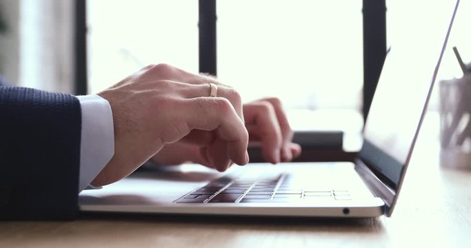 Close up view of male hands using laptop. Businessman wearing suit typing on computer keyboard browsing internet, communicating online, working on modern tech in office. Business software concept.
