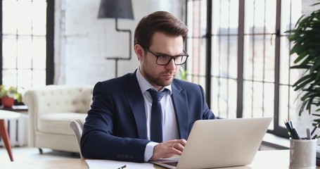 Focused businessman wearing suit doing online research on laptop at work. Serious male executive analyzing digital financial market making notes. Smart stock analyst working on computer in office. - Powered by Adobe