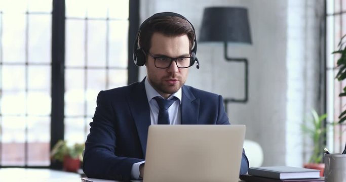 Confident businessman wears suit and headset makes conference video call on laptop. Male call center agent, customer support manager or telemarketer communicating in webcam chat on computer in office.