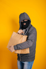 Thief with paper bags. The guy in the balaclava steals purchases from the store. Masked man on yellow background