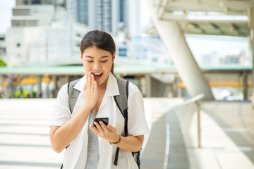 portrait surprised young Asian woman in casual shirt carrying a backpack take her hand close her mouth while watching a cell phone at modern footbridge