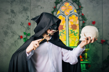 A guy in a black mask in the form of a skull and in a black cloak. Evil sorcerer with a magic wand and skulls in his hand. The wizard in the castle. halloween image