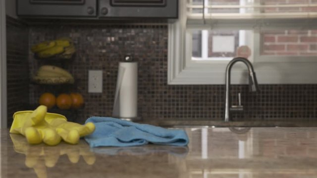 Young man in a gray T-shirt cleaning kitchen granite countertop in his house. He is wearing a bright yellow rubber glove and holds a blue rag which he is using for wiping the kitchen table with a