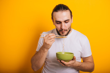 A guy in a white T-shirt stands and eats instant noodles from a green cup on a yellow background. A man dines with noodles using wooden chopsticks. Get pleasure