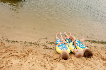 Funny kids in bathing suits sit together on sand with their legs in water. boys are happy on holiday in village together. Summer day, river, swimming in the water. Close-up legs and feet