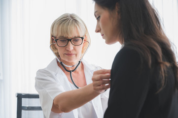 Senior female doctor using stethoscope listening to patients heartbeat in hospital medical office