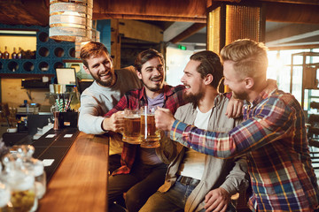 Friends with glasses of beer clinking while standing at a bar in a bar pub.