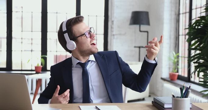 Excited funny businessman wearing suit and wireless headphones listening to rock music sitting at workplace desk. Happy male executive having fun pretending playing guitar relaxing in office at break.