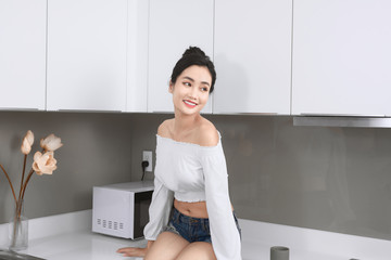 Cheerful woman sits on countertop in white modern kitchen. Morning time. Home clothes.