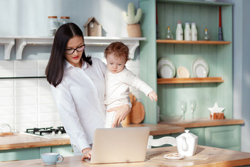 Obraz premium Young mother works at home with a laptop with a baby in her arms, remote work on maternity leave