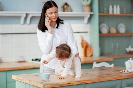 Young mother talking on the phone at home in the kitchen with a baby in her arms, remote work on maternity leave