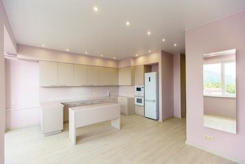 A large room with pink walls and a white kitchen set. Kitchen furniture is new with all kitchen appliances. In front of the kitchen is a white table. Fresh, new renovation.