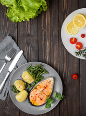 Cooked salmon steak with potatoes and green beans on a plate, dark wooden vintage background. Nearby fresh vegetables. Top view, lunch concept or menu of red fish.