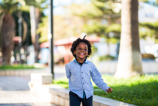 Happy And Beautiful Young Kid Black Race African With Ethnic Alternative Hair Playing And Runnig  In A Playground With  And Have Fun - City Street In Background