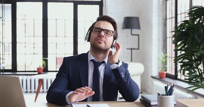 Relaxed funny male executive listening to music wearing wireless headphones in office. Carefree happy businessman in suit enjoying stress free break sitting at work desk feeling peace of mind concept.