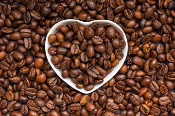 White porcelain heart filled with dark roasted coffee beans on a background with brown coffee beans. Symmetrical image.