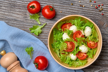 Fresh salad with cherry tomatoes, mozzarella cheese, greens leaves in bowl. Healthy summer salad vegetarian meal concept. Tomatoes and lettuce home made caprese salad on wood, top view