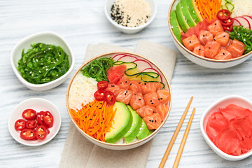 Hawaii salmon poke, avocado, rice, seaweed, cucumber, radish, carrot in bowl. Home made vegetarian poke. Seafood concept. Asian salmon poke salad on white wood background.