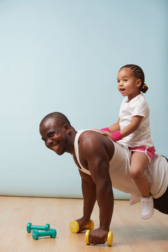 Handsome black young father is doing push ups on dumbbells with his cute little daughter riding him. They both enjoy it big time. Indoors.
