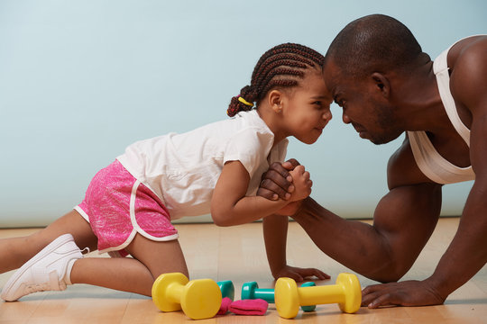 Handsome Black Young Father Is Arm Wrestling With His Cute Little Daughter On The Floor At Home. They Looking At Each Other Angrily, Trying To Show Their Resolve. Grimacing At Each Other.