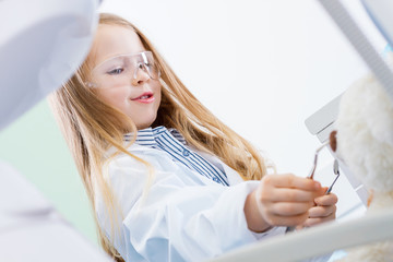 Little cute girl in white doctor robe uniform and protective glasses is treating with instruments, tools her patient bear toy in chair. Child is playing in dentist in office, clinic. Dentistry concept