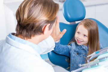 Little cute girl is sitting in dental chair in clinic, office. Friendly man doctor is giving kid five. Orthodontist is preparing for examination of patient. Visiting dentist with children.