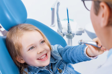 Little girl is sitting in dental chair, holding in hand tool. Orthodontist is preparing for examination child teeth. Friendly relationships between doctor and patient. Visiting dentist with children.