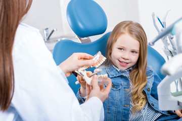 Obraz na płótnie Canvas Girl is sitting in dental chair in clinic, office. Woman doctor is showing jaw layout, teaching how brush teeth. Orthodontist is preparing for examination of patient. Visiting dentist with children.