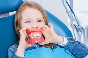 Little cute girl is sitting in dental blue chair in clinic, office. Funny kid patient is holding...