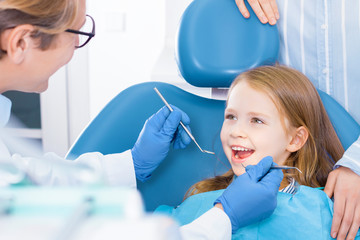Little cute smiling girl is sitting in dental chair and opening mouth in clinic, office. Doctor is preparing for examination of child teeth with tools, instruments. Visiting dentist with children.