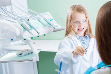 Little cute girl in white doctors coat and protective glasses is treating with instruments, tools teeth of woman patient in chair. Child is playing in dentist, orthodontist. Dentistry concept.