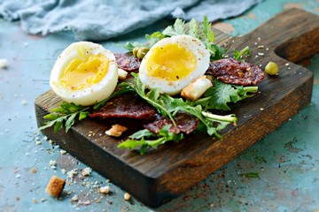 Soft-boiled eggs for Breakfast with chorizo sausage, fresh arugula salad and croutons (crackers) on a wooden board on a blue background.