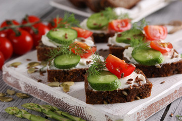 Healthy food. Sliced dark bread with cream cheese, cucumber, red cherry tomatoes and dill on a white wooden board. Pumpkin, flax seeds, lettuce, asparagus on a light wooden background. Copy space