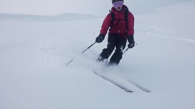 Slow motion shot of a man wearing red jacket, skiing downhill, surrounded by snow white mountain and semi sunny background covered with clouds.