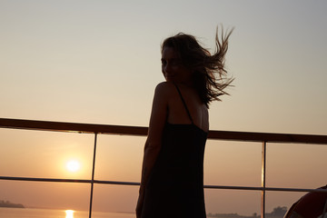 Fototapeta premium Sensual authentic shot of attractive young Caucasian woman in long black dress, posing on board of cruise liner, feeling happy and confident, enjoying picturesque sunset with great river view.