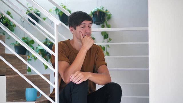 Pensive sad and depressed young man sitting alone on stairs at home: painful sad man.