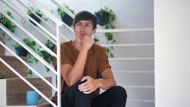 Pensive sad and depressed young man sitting alone on stairs at home: painful sad man.
