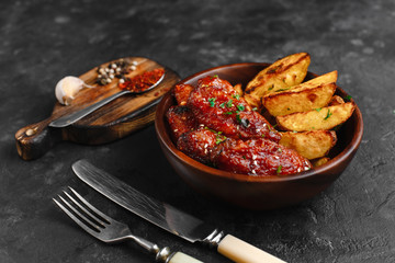 Grilled chicken wings in barbecue sauce with sesame and parsley, with rustic potatoes in a wooden plate on a dark table. Top view with space for text. Delicious beer appetizer on a dark background.