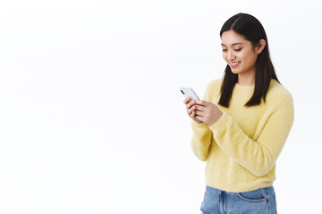 Fototapeta premium Happy cute asian girl using mobile phone and smiling. Female student sending funny meme via social media messanger, chat with friends or team members, video-call on smartphone, white background