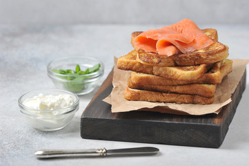 Slices of fried bread and slices of salmon with weak salt. Next to toast is a knife and cups with herbs and soft cheese. Light background. Close-up.