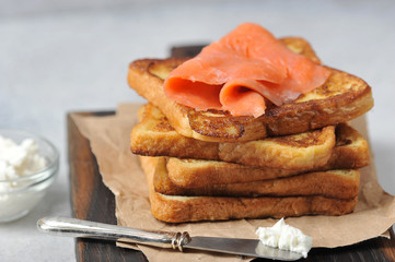 Slices of fried bread and slices of salmon with weak salt. Next to a knife and a cup with soft cheese. Light background. Close-up. 