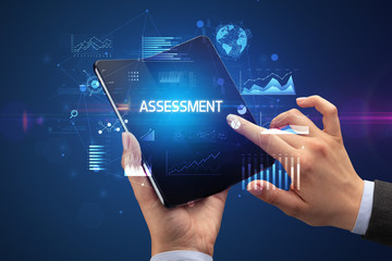 Businessman holding a foldable smartphone with ASSESSMENT inscription, successful business concept