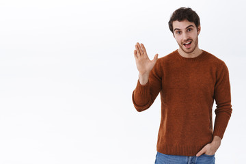 Fototapeta premium Hi there. Handsome caucasian 25s guy in sweatshirt, hold hand in pocket and casually informal greet person, smiling and waving raised hand in hello gesture, welcome, introduce himself